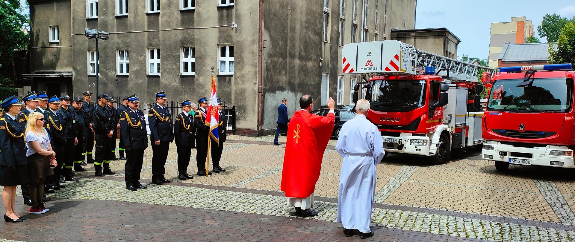 Zdjęcie przedstawia księdza, który odprawia poświęcenie pojazdów pożarniczych znajdujących się przed budynkiem kościoła.