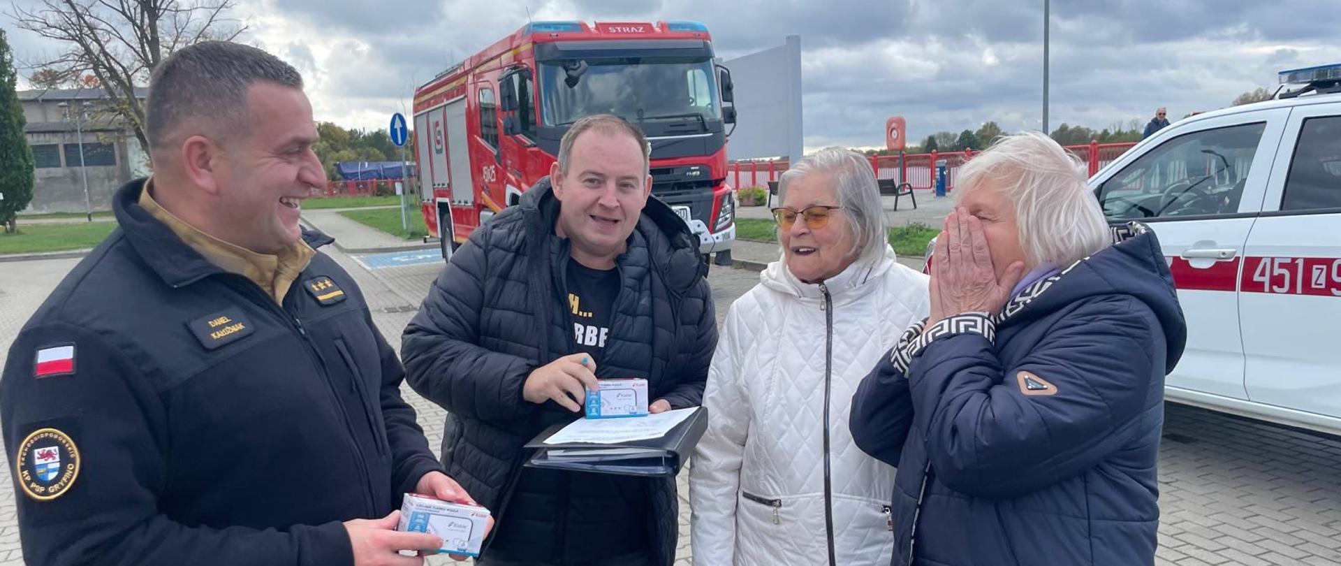 Przekazywanie czujek mieszkańcom przez Zastępcę Komendanta Powiatowego Państwowej Straży Pożarnej w Gryfinie i pracownika Urzędu Miasta i Gminy Gryfino. W tle pojazdy pożarnicze - jeden biały pickup i jeden czerwony pojazd ciężarowy.