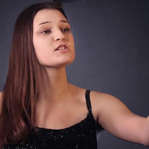 Zdjęcie kolorowe. Dziewczyna w stroju wieczorowym. Śpiewa. Stoi z wyciągniętą w przód lewą ręką.