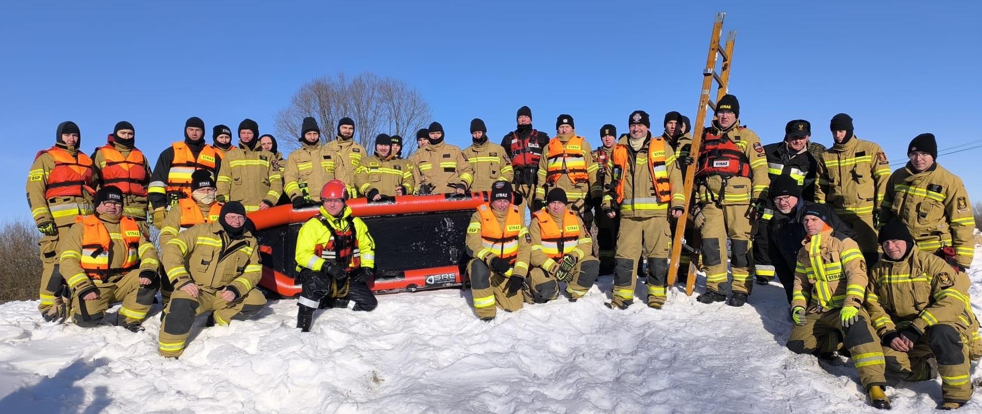 Na śnieżnym terenie stoi duża grupa strażaków pozujących do wspólnego zdjęcia. Wszyscy mają na sobie pełne umundurowanie ochronne, a część dodatkowo kamizelki asekuracyjne w jaskrawych kolorach. W centrum ustawiono dmuchaną łódź ratowniczą, a za nią oparta jest pionowo drewniana drabina. W tle widać czyste, błękitne niebo, co nadaje zdjęciu spokojny, zimowy charakter. Całość wygląda jak pamiątkowa fotografia z ćwiczeń lub szkolenia w warunkach zimowych.