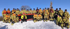 Na śnieżnym terenie stoi duża grupa strażaków pozujących do wspólnego zdjęcia. Wszyscy mają na sobie pełne umundurowanie ochronne, a część dodatkowo kamizelki asekuracyjne w jaskrawych kolorach. W centrum ustawiono dmuchaną łódź ratowniczą, a za nią oparta jest pionowo drewniana drabina. W tle widać czyste, błękitne niebo, co nadaje zdjęciu spokojny, zimowy charakter. Całość wygląda jak pamiątkowa fotografia z ćwiczeń lub szkolenia w warunkach zimowych.