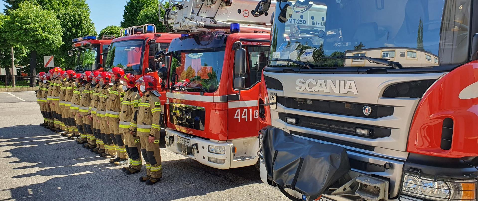 Zdjęcie przedstawia czerwone samochody gaśnicze, przed którymi stoją strażacy w piaskowych mundurach specjalnych i czerwonych hełmach. W tle zielone korony drzew.