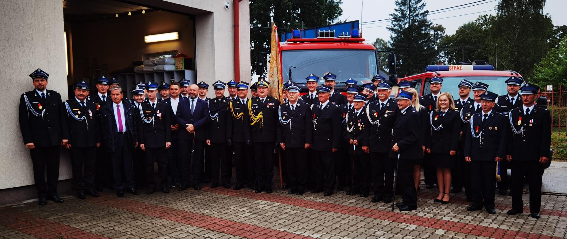 strażacy psp, druhowie osp inni uczestnicy uroczystości na zbiorowym zdjęciu, za zgromadzonymi budynek remizy oraz samochody pożarnicze