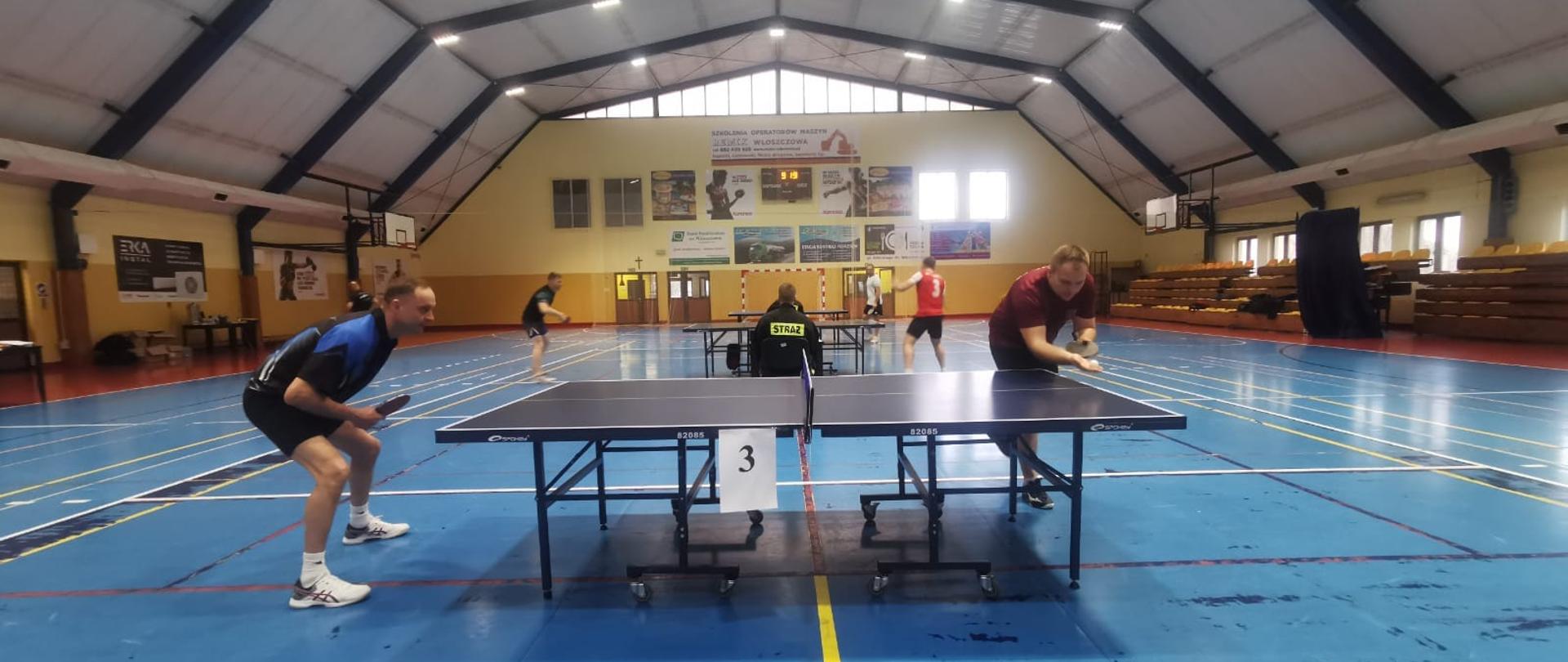 Na zdjęciu widzimy mecz w tenisa stołowego. Rywalizuje dwóch zawodników. W tle drugi pojedynek.