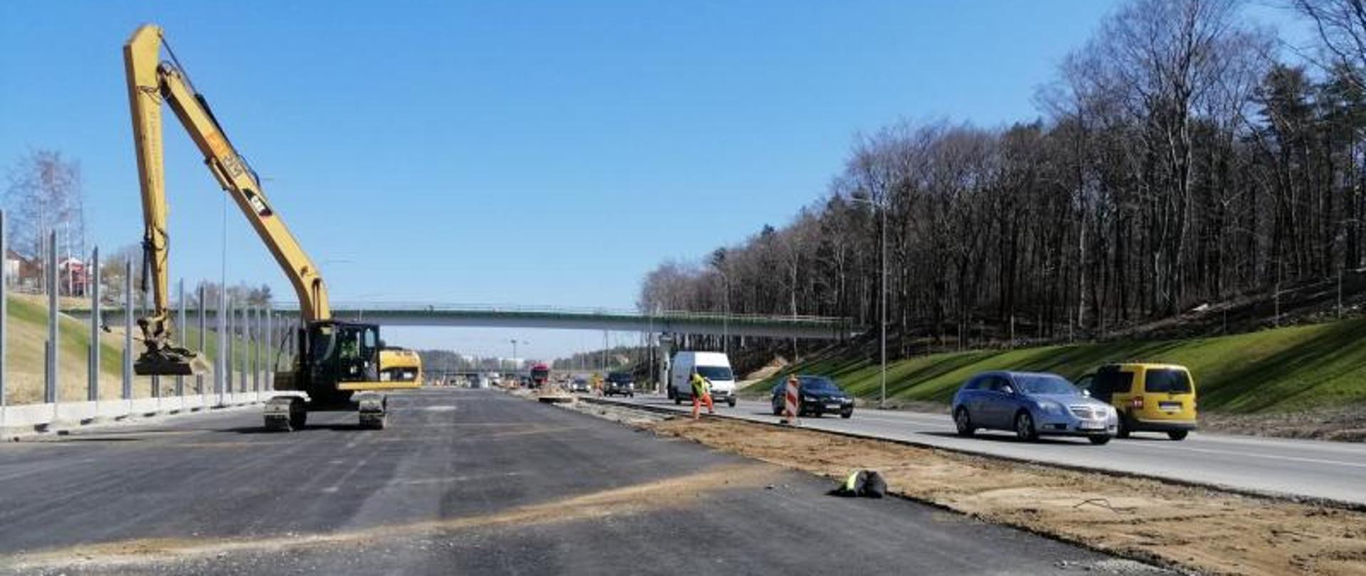 Budowa drogi S6 w Gdyni. Zdjęcie przedstawia nową nawierzchnię przyszłej drogi ekspresowej i pracującą koparkę. Po prawej odbywa się ruch samochodów.