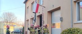 Po prawej stronie przy budynku stoi kilku strażaków, przed nimi poczet flagowy wciąga flagę Polski na maszt. Przed masztem zielone przycięte krzewy. Po lewej widoczny asfaltowy plac i chodnik .