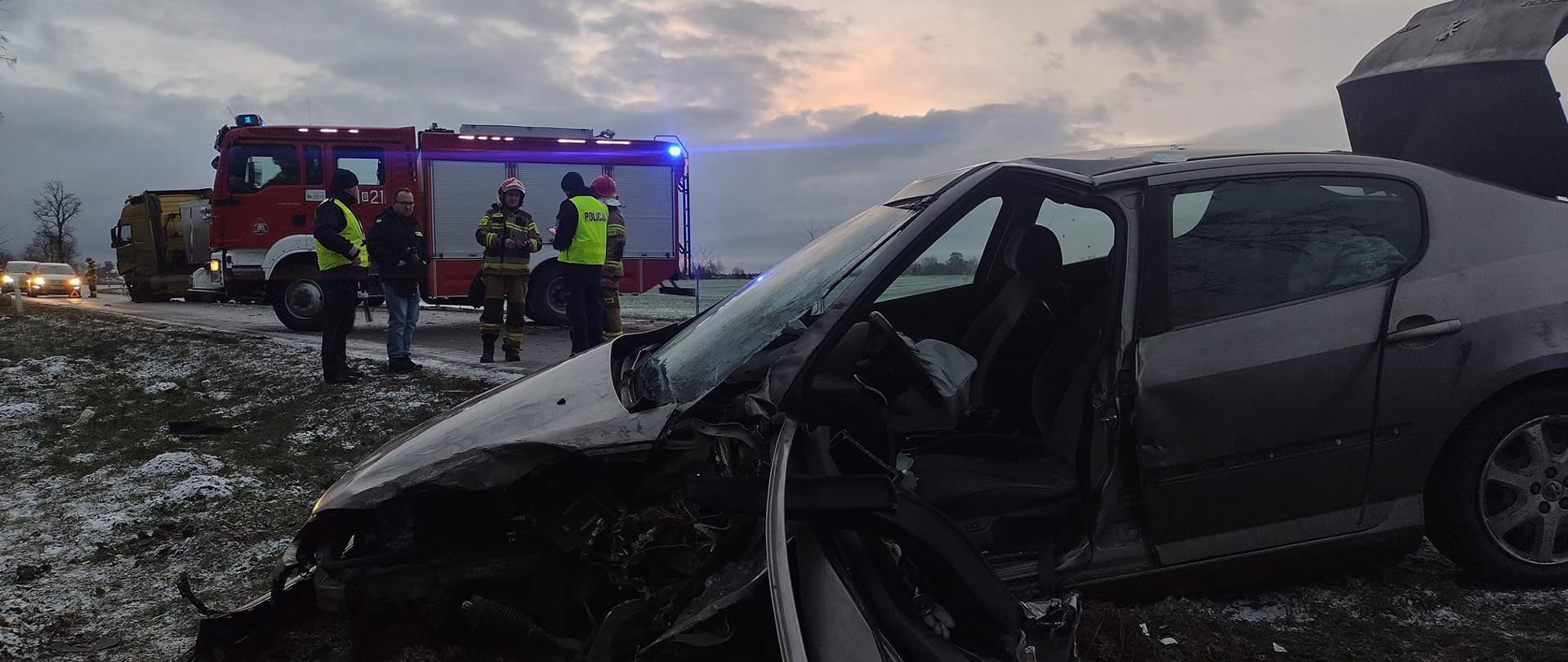 Uszkodzony samochód osobowy na pierwszym planie, w tle samochód straży pożarnej