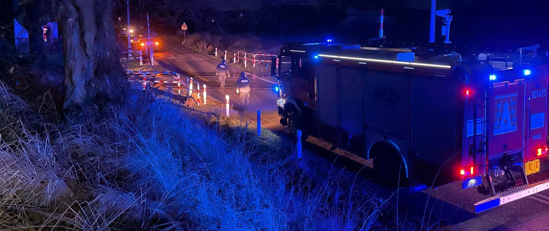 Zdjęcie przedstawia scenę nocnej interwencji służb ratunkowych na drodze, oświetloną głównie przez niebieskie światła awaryjne pojazdów. Na pierwszym planie po prawej stronie znajduje się tył wozu strażackiego, z włączonymi niebieskimi światłami sygnalizacyjnymi, które rzucają intensywny niebieski blask na otoczenie. Na tylnej części wozu widoczny jest numer "601 25". Droga w oddali jest częściowo zablokowana przez barierki odblaskowe i pachołki, co wskazuje na zabezpieczenie miejsca zdarzenia. Wzdłuż drogi widać rozstawione lampy ostrzegawcze, emitujące pomarańczowe światło. Po lewej stronie drogi, w oddali, idą dwie osoby w ciemnych, prawdopodobnie funkcjonariusze Policji. Tło zdjęcia stanowią ciemne drzewa i krzewy. Światła latarni ulicznych w oddali również oświetlają scenę. Ciemne niebo potęguje wrażenie nocy. Całość obrazu wskazuje na akcję ratowniczą prowadzoną w ciemności.
