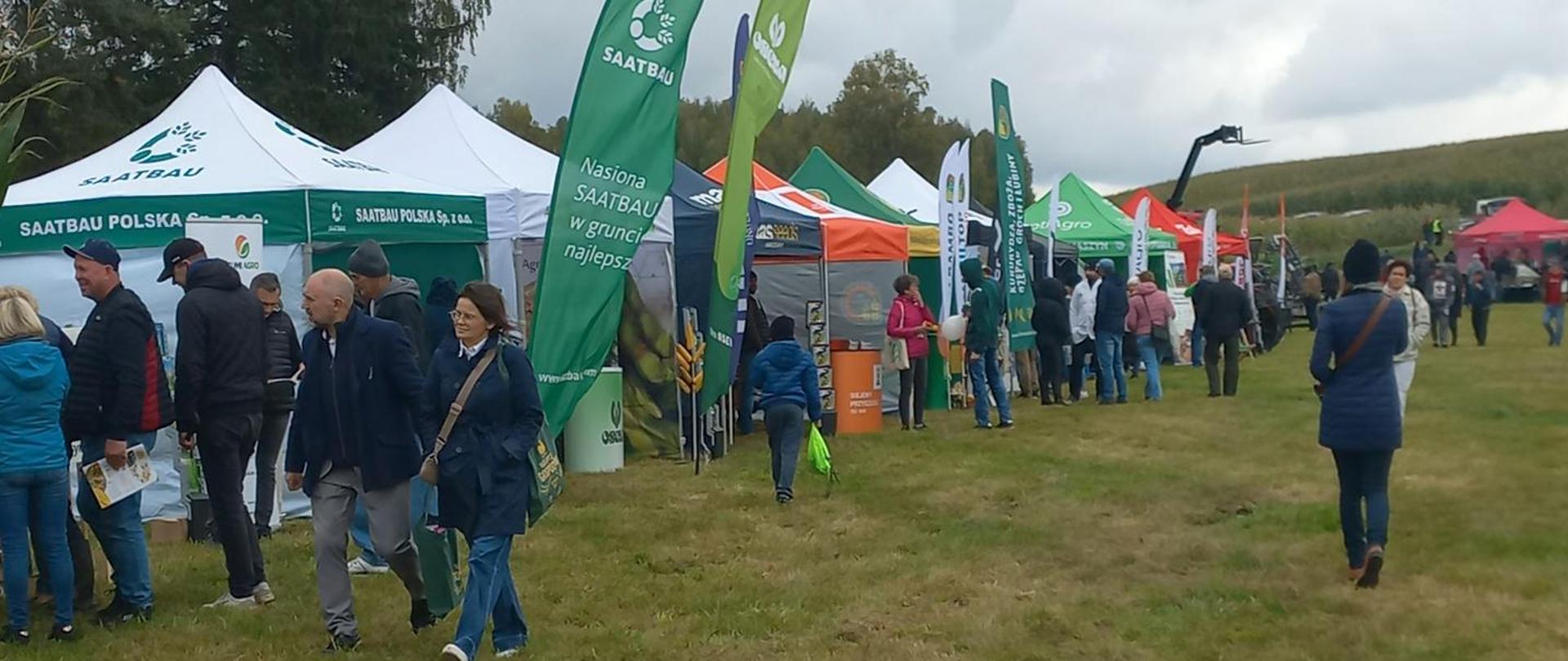 Widok na alejkę targową w terenie, gdzie wzdłuż stoisk z namiotami i flagami firm spacerują uczestnicy wydarzenia.