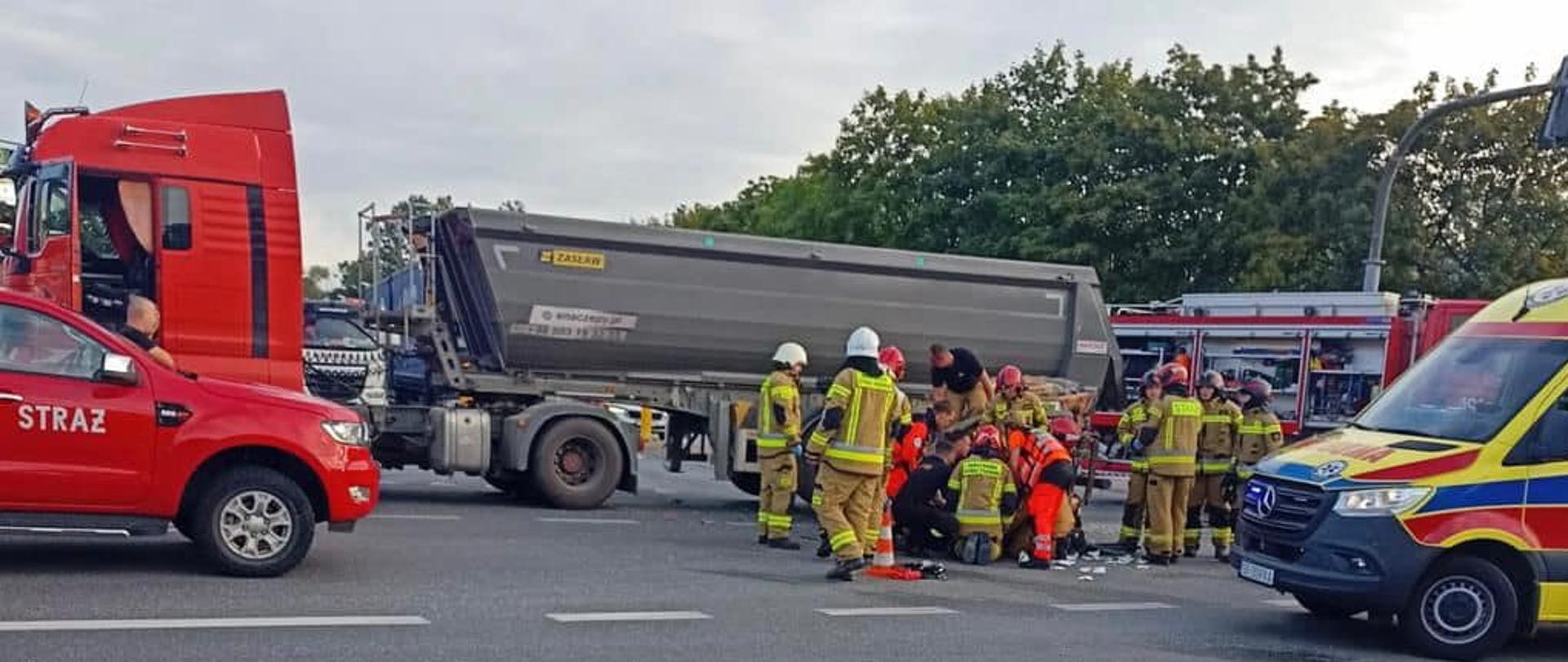Na zdjęciu znajduje się skrzyżowanie na którym stoją pojazdy strażackie, ZRM, naczepa ciężarówki. Po skrzyżowaniu chodzą strażacy i "medycy"