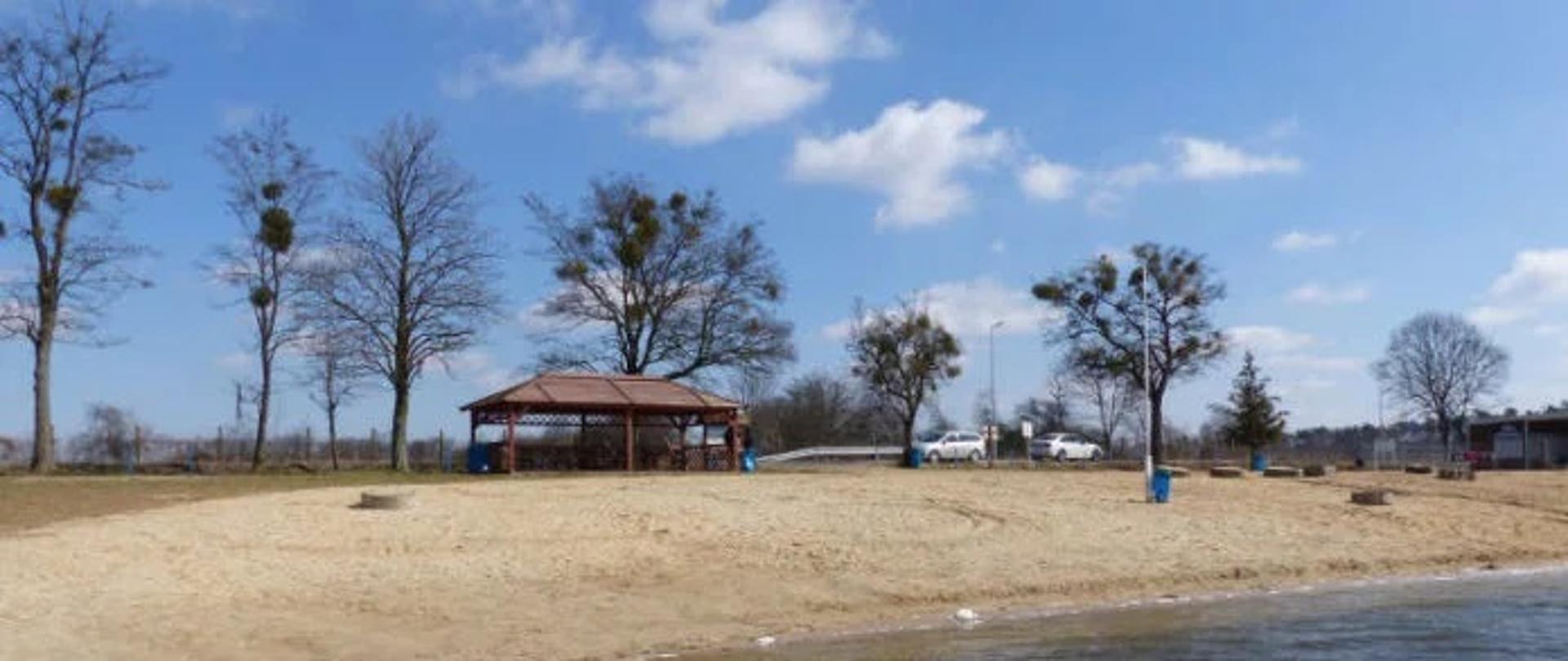 Widać wiatę z drewna na plaży, za nią kilka drzew oraz brzeg jeziora