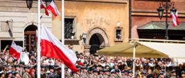 Na zdjęciu Plac Zamkowy w Warszawie podczas uroczystości obchodów 235. rocznicy uchwalenia Konstytucji 3 Maja. Na zdjęciu po prawej stronie orkiestra wojskowa, w środku maszt, na którą żołnierze wciągają biało-czerwoną flagę Polski. Po lewej stronie od masztu kompania honorowa Marynarki Wojennej. W tle widać widzów oraz kamienice starówki. Na latarniach małe flagi biało-czerwone. 