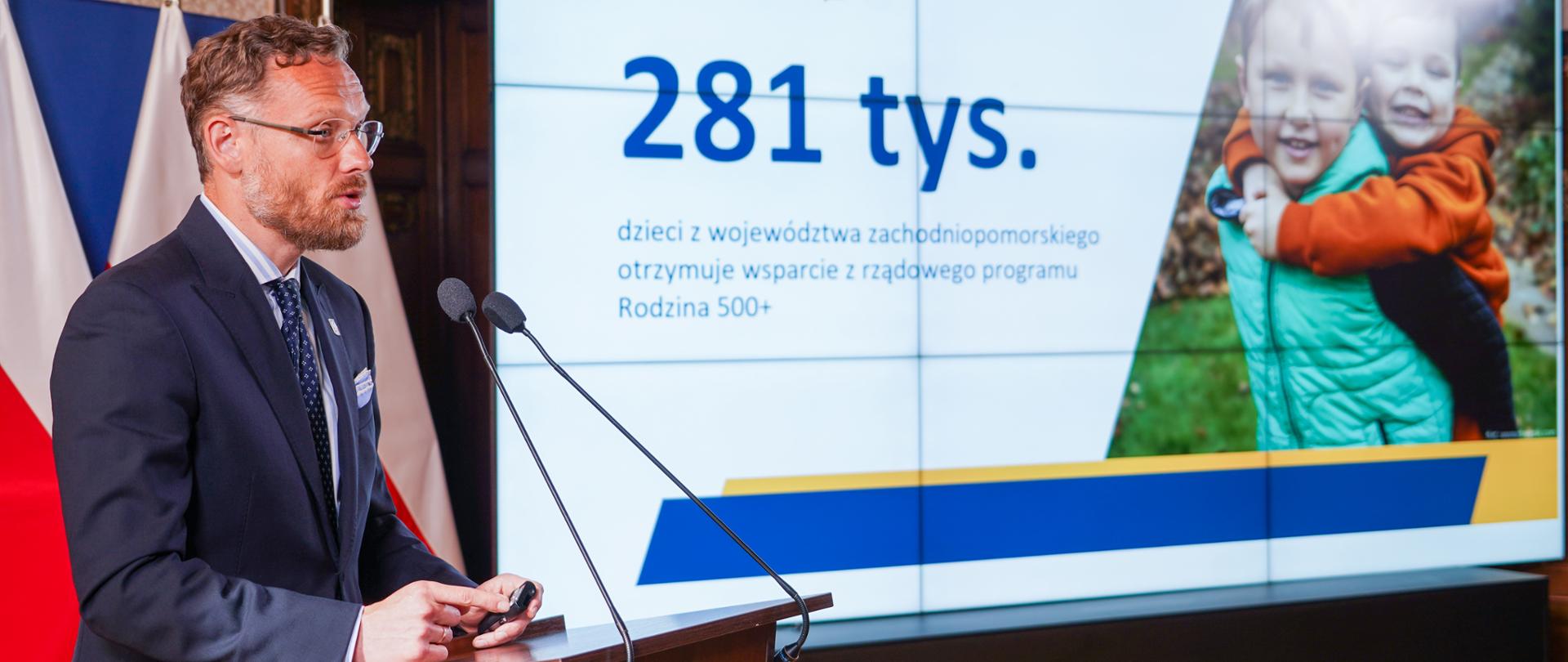 wojewoda Zbigniew Bogucki przy mównicy, w tle liczba: 281 tys. dzieci objętych programem Rodzina 500 Plus