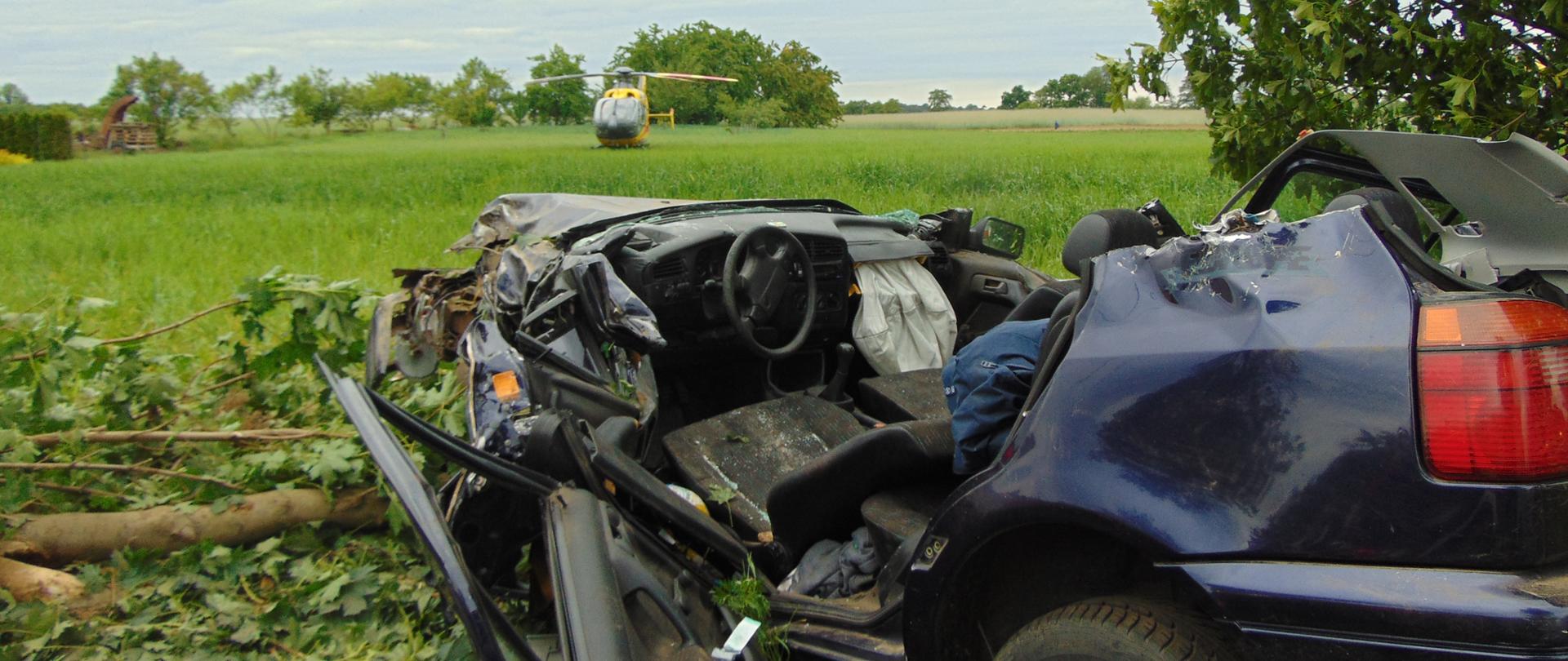 Samochód stoi na poboczu drogi przy drzewie, w które uderzył. Pojazd ma odcięty dach oraz drzwi po stronie kierowcy. W tle na polu widać stojący śmigłowiec LPR.