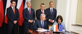 przy stoliku z flagami polską i unijną dwie osoby podpisują umowę za nimi stoją pozostali uczestnicy spotkania