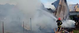 Zdjęcie przedstawia strażaka prowadzącego działania gaśnicze podczas pożaru budynku gospodarczego wraz z przylegającą bezpośrednio do niego wiatą z drewnem