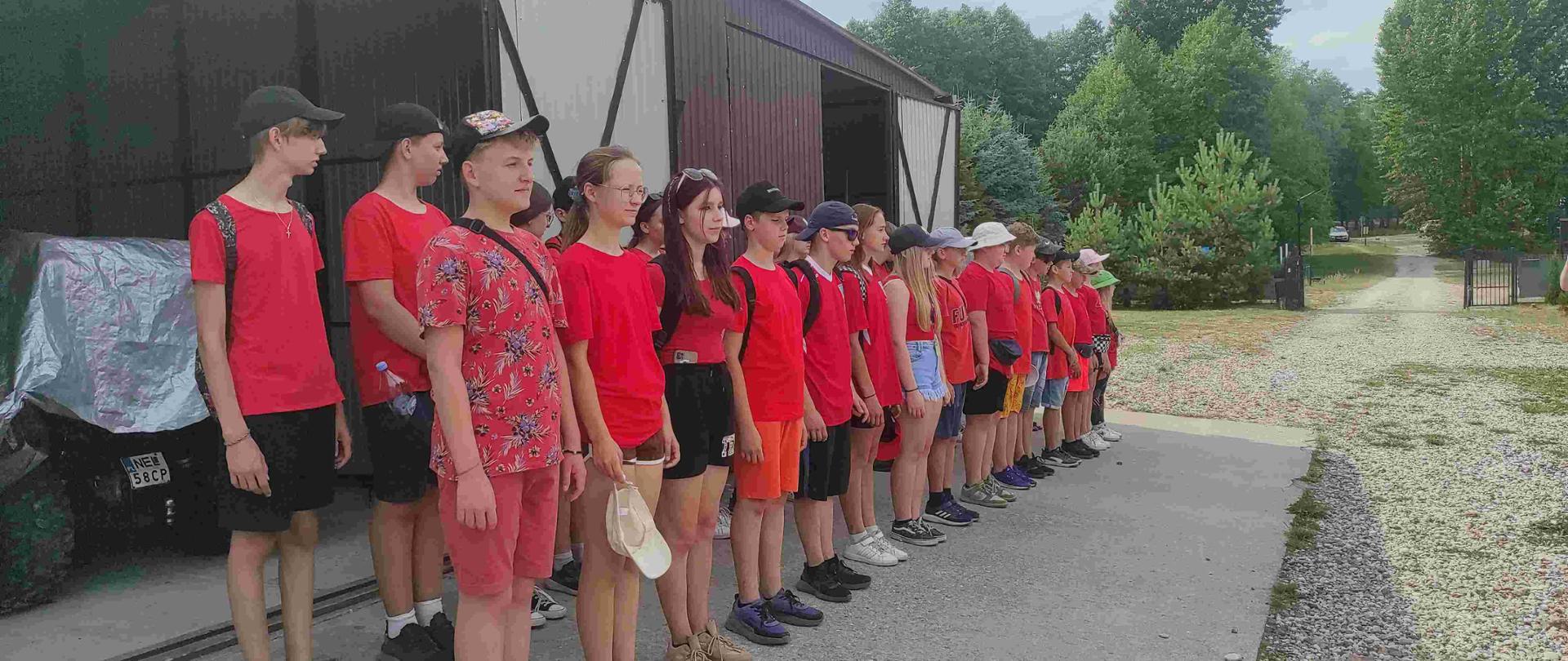 Przed garażem stoi kilkadziesiąt dzieci w czerwonych koszulkach w dwuszeregu.