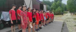 Przed garażem stoi kilkadziesiąt dzieci w czerwonych koszulkach w dwuszeregu.