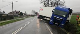 Zdjęcie przedstawia samochód ciężarowy z naczepą w rowie. Tył naczepy blokuje jeden pas drogi. W tle dwóch strażaków PSP wykonujących zlecone czynności.