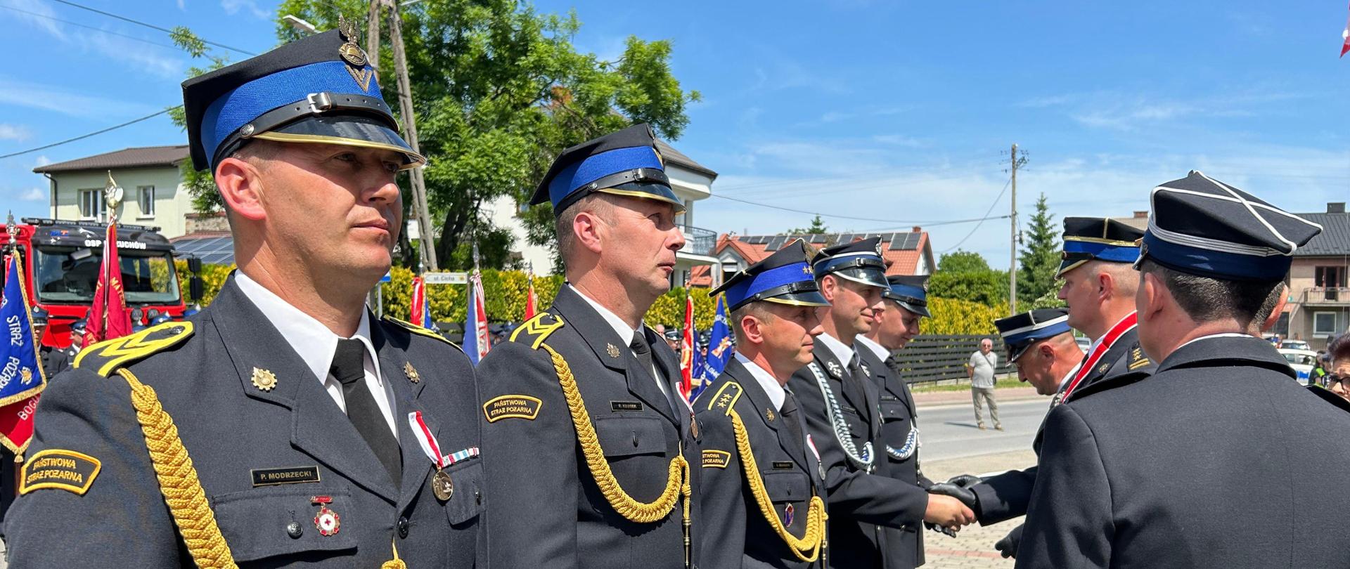 Grupa strażaków w galowych mundurach stoi w rzędzie na zewnątrz. Dwóch mężczyzn na pierwszym planie ma na sobie granatowe mundury ze złotymi sznurami naramiennymi i granatowe czapki z dystynkcjami. W tle widać budynki, drzewa i flagi. Czwarty strażak od lewej przyjmuje jakieś odznaczenie i gratulacje od oficera Państwowej Straży Pożarnej w stopniu starszego brygadiera. W tle widać budynki, linię energetyczną i osobę cywilną.