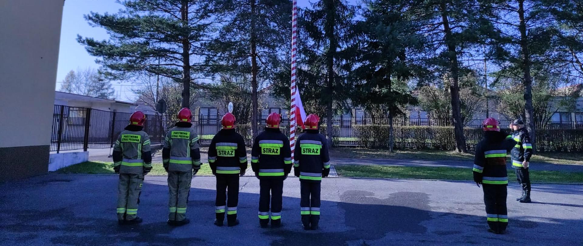 Na zdjęciu widzimy strażaków podczas uroczystej zbiórki z okazji Święta Niepodległości. Trwa wciąganie flagi na maszt i śpiew Hymnu. 