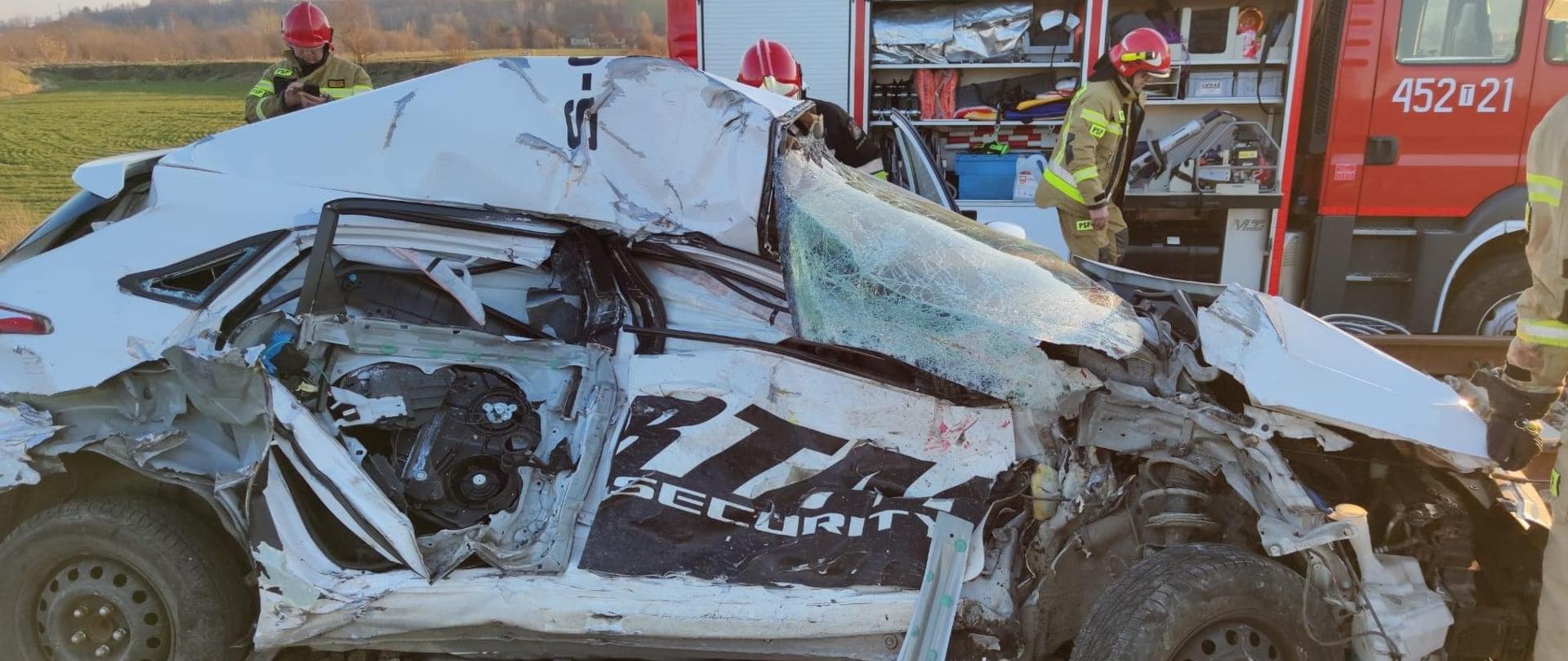 Zmiażdżony samochód osobowy - pogięta karoseria znajdujący się na torach kolejowych. Za autem samochód gaśniczy oraz strażacy zabezpieczający teren zdarzenia. 