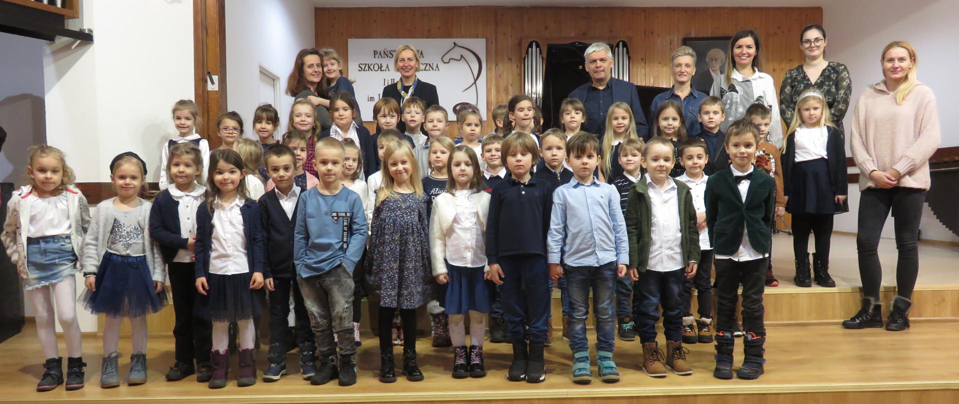 Dzieci stoją na scenie sali koncertowej PSMw 4 rzędach, z tyłu opiekunowie i organizatorzy. W tle logo PSM