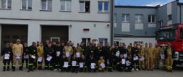 Zdjęcie zbiorowe uczestników szkolenia podstawowego strażaków ratowników Ochotniczych Straży Pożarnych. Obok uczestników trzymających w dłoniach dyplomy stoją funkcjonariusze Komendy. Po prawo widoczny jest przód samochodu gaśniczego. W tle budynek Jednostki Ratowniczo Gaśniczej.