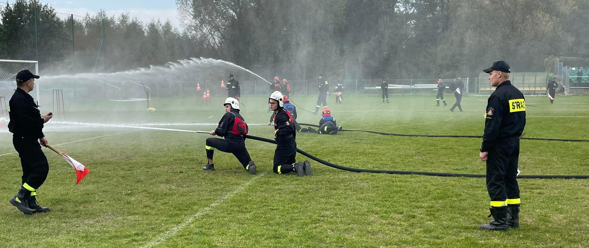 dwóch strażaków ochotników podaje przed siebie zwarty prąd wody. Obok nich stoi dwóch sędziów.
