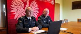 Komendanci siedzą przy stole w sali konferencyjnej w umundurowaniu wyjściowym. Na stole znajduje się laptop, dokumenty, piloty oraz telefon. W tle baner Państwowej Straży Pożarnej w kolorze czerwonym.