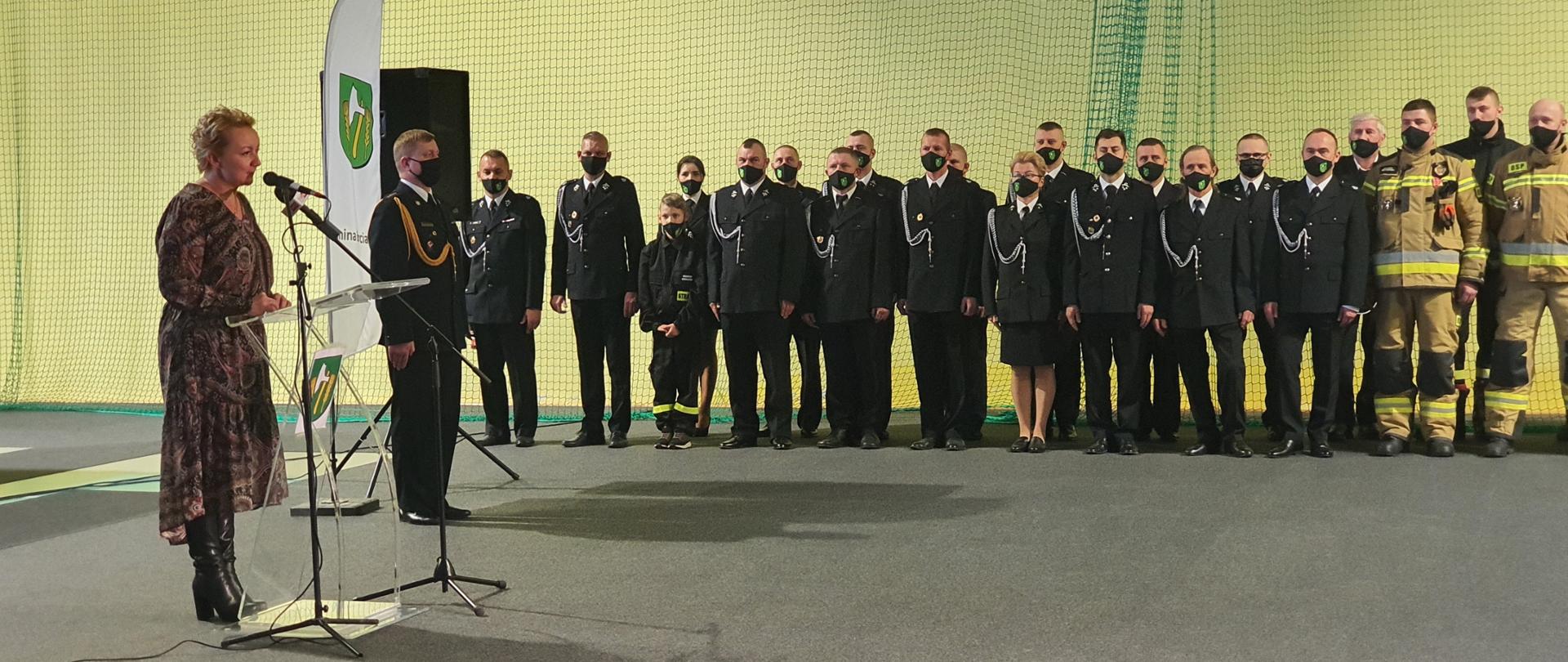 Zbiórka z okazji przekazania sprzętu dla OSP. Na zdjęciu widać kobietę przemawiającą do zebranych oraz druhów w szyku