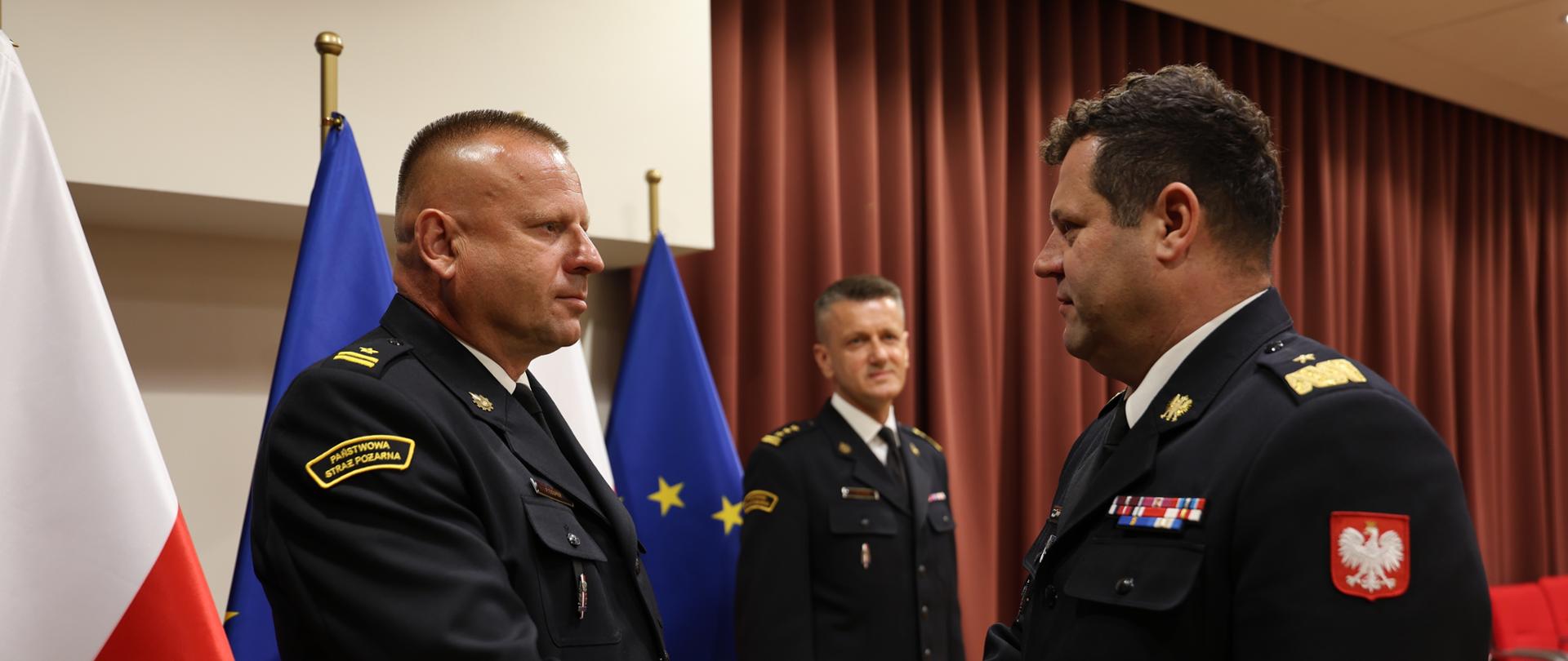 Komendant wojewódzki ściska dłoń nowo powołanemu Zastępcy Komendanta. Mężczyźni ubrani w strażackie mundury wyjściowe znajdują się na sali konferencyjnej. W tle widoczny Komendant Powiatowy PSP w Wyszkowie.
