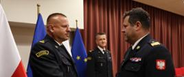 Komendant wojewódzki ściska dłoń nowo powołanemu Zastępcy Komendanta. Mężczyźni ubrani w strażackie mundury wyjściowe znajdują się na sali konferencyjnej. W tle widoczny Komendant Powiatowy PSP w Wyszkowie.