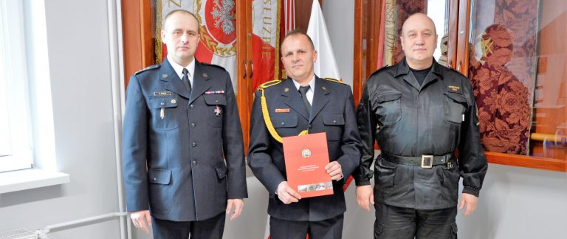 Na pamiątkowym zdjęciu Komendant Powiatowy wraz z zastępcą i Krzysztofem Orłowskim. Komendant oraz Krzysztof Orłowski w mundurach galowych. Zastępca w mundurze służbowym. W tle witryny ze sztandarem oraz biało czerwona flaga.