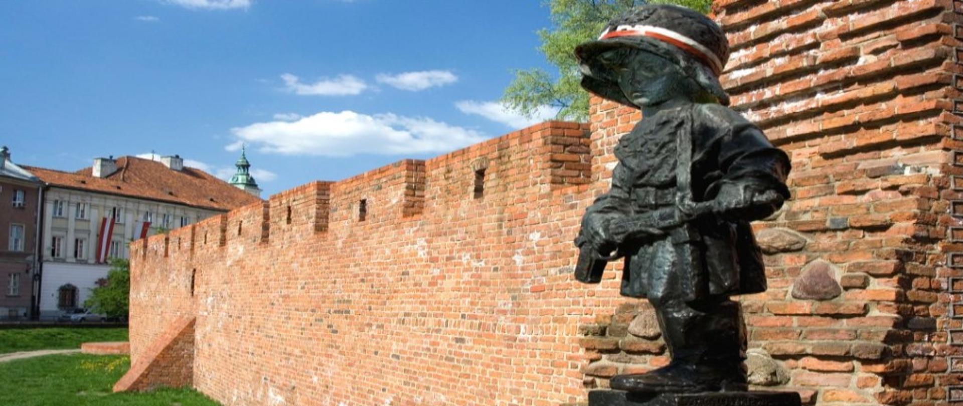 Rzeźba małego dziecka z karabinem w rękach i w hełmie, w tle stary mur obronny