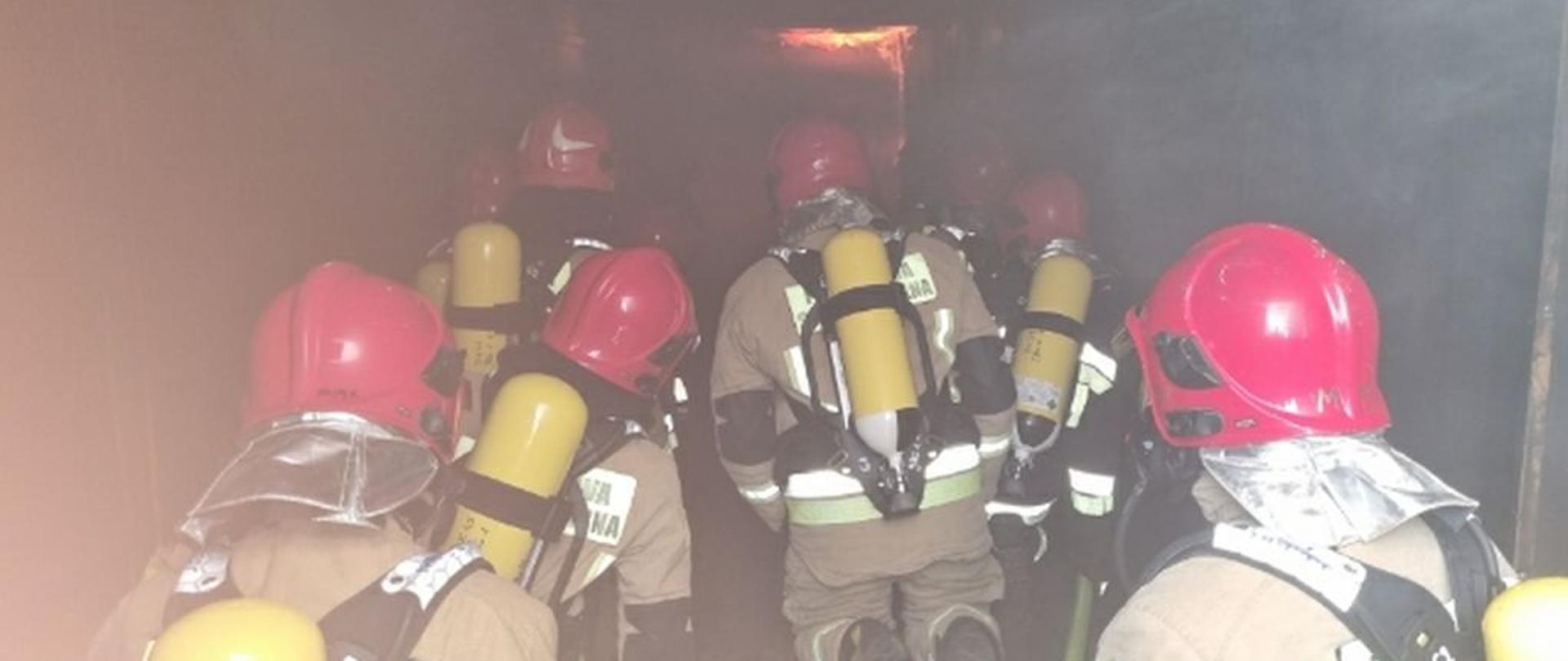 Zdjęcie przedstawia strażaków wewnątrz trenażera. Strażacy są w pozycji klęczącej. ubrani są w odzież specjalną i zabezpieczeni są sprzętem ochrony dróg oddechowych. Nad ich głowami zabezpieczonymi w hełm snują się kłęby dymu. W dali widać języki ognia.