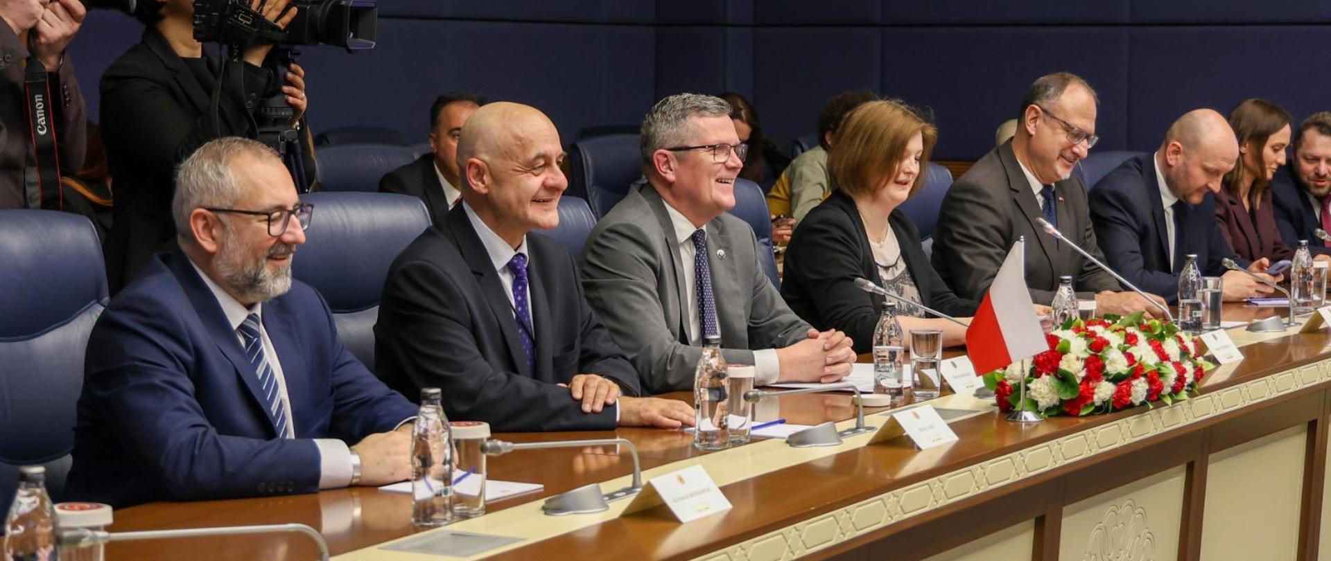 Członkowie delegacji – pięciu mężczyzn i dwie kobiety – siedzą przy stole konferencyjnym podczas rozmów bilateralnych. Uczestnicy ubrani są w formalne stroje biznesowe: mężczyźni w garniturach i krawatach, kobiety w marynarkach lub eleganckich bluzkach.