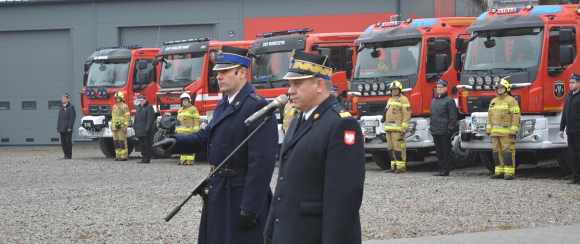 Zdjęcie przestawia Mazowieckiego Komendanta Wojewódzkiego Państwowej Straży Pożarnej nadbrygadiera Jarosława Nowosielskiego stojącego w asyście drugiego funkcjonariusza Państwowej Straży Pożarnej pełniącego funkcję dowódcy uroczystości. Funkcjonariusze ubrani są w płaszcze oraz mundury galowe. Komendant stoi przed mikrofonem. W tle za funkcjonariuszami widać ustawione pojazdy pożarnicze. Pojazdy w kolorze czerwonym z białymi i srebrnymi elementami zabudowy. Przy pojazdach ustawieni są inni strażacy, ubrani w kurtki służbowe oraz mundury galowe. Kierowcy pojazdów ubrani są w ubrania specjalne w kolorze piaskowym. Na głowach mają hełmy. Strażacy ci będą odbierać w trakcie apelu kluczyki do nowo przekazywanych pojazdów.