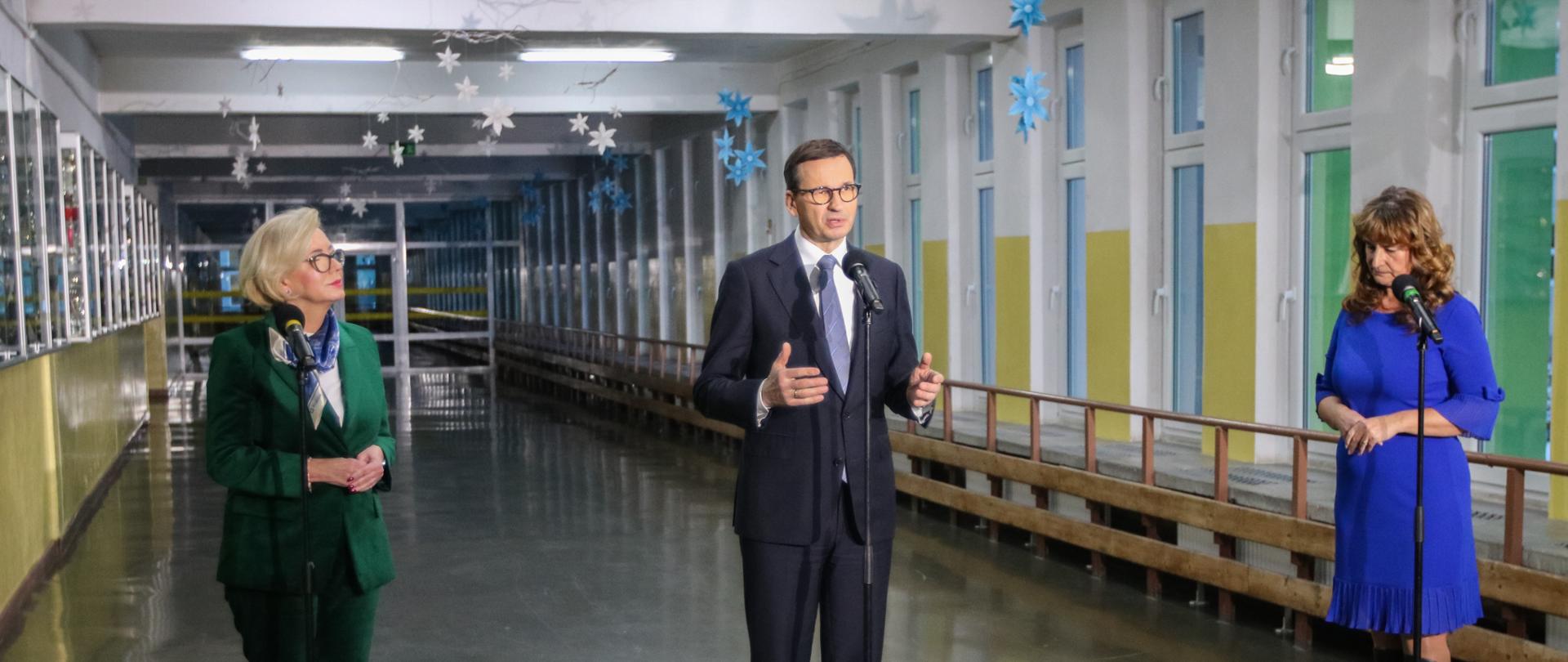 Na szkolnym korytarzu na błyszczącej podłodze stoją trzy osoby przy trzech mikrofonach - wiceminister Machałek, premier Morawiecki i kobieta ubrana na niebiesko.