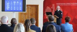 Komendant powiatowy przemawia z mównicy. Na pierwszym planie słuchacze podsumowania. W tle po prawej stronie baner reklamowy Komendy Powiatowej Państwowej Straży Pożarnej w Ostrzeszowie; po lewej stronie monitor z statystykami podejmowanych interwencji w 2023 roku.