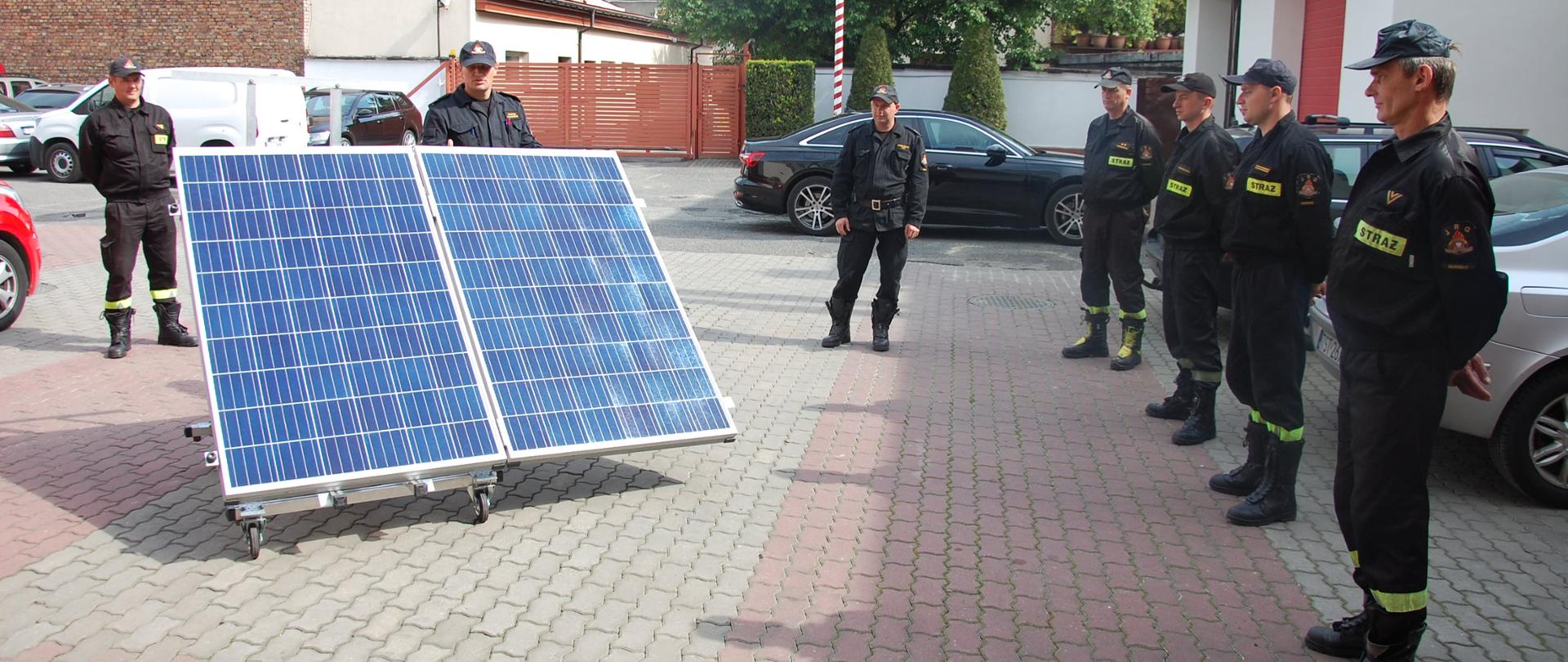Z lewej strony zdjęcia widać dwa panele fotowoltaiczne skierowane szybą w naszym kierunku. Za panelami stoi mężczyzna, który jest instruktorem, oraz mężczyzna, strażak, który jest uczestnikiem szkolenia. Po prawej stronie zdjęcia widać 5 mężczyzn wśród których stoi kolejny instruktor. Wszyscy mężczyźni stoją na dworze, mają na sobie czarne ubrania a na głowach mają czarne czapki z daszkiem. W tle zdjęcia widać samochody osobowe oraz budynki. Plac na którym stoją ludzie jest wyłożony szarą i czerwoną kostką brukową. 