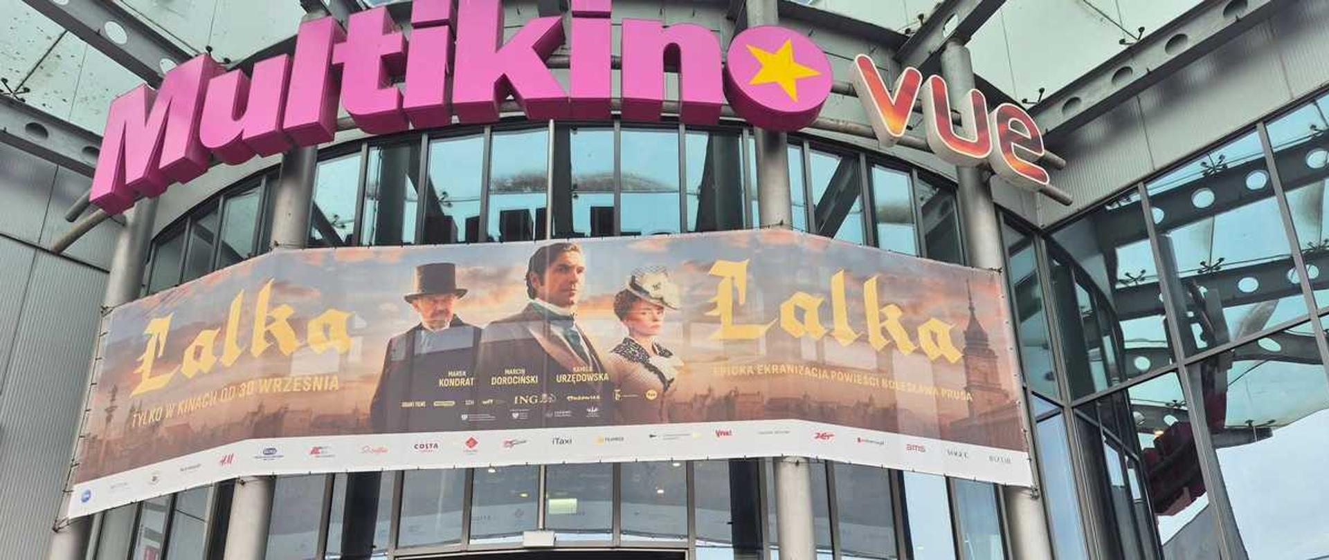"Na zdjęciu widoczna jest grupa około dwudziestu nastolatków wraz z opiekunem, stojących przed wejściem do kina Multikino VUE. Grupa pozuje do wspólnego zdjęcia na schodach prowadzących do przeszklonych drzwi obrotowych.
Nad wejściem dominuje wielki, różowy szyld z napisem „Multikino”, uzupełniony mniejszym logo „VUE” z żółtą gwiazdą. Bezpośrednio pod nim rozciąga się ogromny, panoramiczny baner reklamowy filmu „Lalka” [tv-movie]. Plakat utrzymany jest w ciepłej, sepiowej tonacji i przedstawia trójkę głównych bohaterów w strojach z epoki. Po lewej i prawej stronie plakatu widnieje duży, ozdobny napis „Lalka”, a po lewej stronie mniejszy tekst: „TYLKO W KINACH OD 30 WRZEŚNIA” [tv-movie]. Na dole baneru umieszczono biały pasek z logotypami sponsorów i partnerów.
Większość grupy stanowią młodzi chłopcy ubrani w luźne, sportowe i codzienne stroje – bluzy z kapturem (w tym jedna czarna marki Adidas), t-shirty i jeansy. Wśród nich wyróżnia się jedna dziewczyna w ciemnej bluzie oraz mężczyzna w średnim wieku (prawdopodobnie nauczyciel lub opiekun) w zielonej koszulce polo i z przewieszoną przez ramię czarną torbą. Kilku chłopców w środku stawki trzyma w rękach kinowe przekąski: duże kubełki z popcornem oraz napoje w kubkach ze słomką.
Konstrukcja budynku jest nowoczesna, wykonana z metalowych elementów i dużych tafli szkła, w których odbija się jasne niebo z delikatnymi chmurami. Po obu stronach schodów zamontowane są solidne, metalowe barierki ochronne. Cała scena jest dobrze doświetlona naturalnym światłem dziennym."