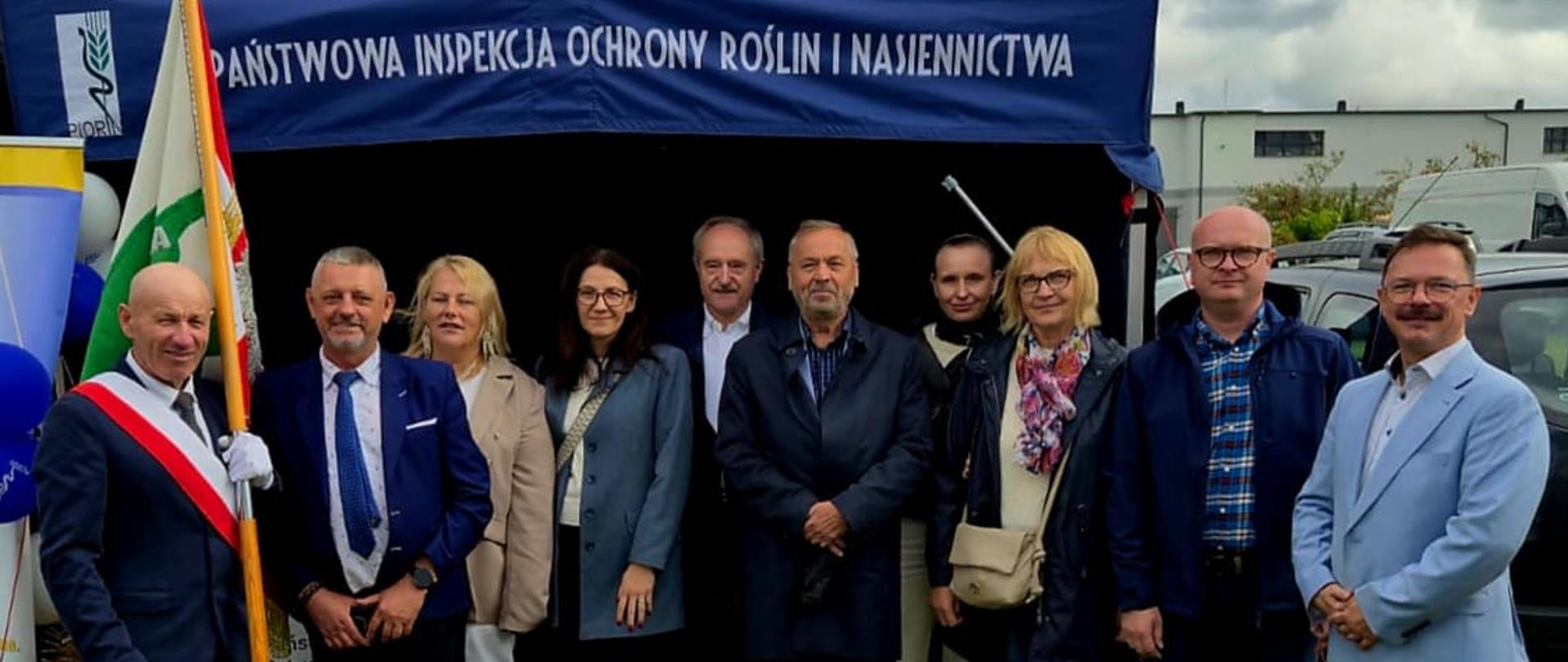 Grupa ludzi, w tym jedna osoba ze sztandarem, stojąca przed granatowym namiotem 