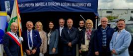 Grupa ludzi, w tym jedna osoba ze sztandarem, stojąca przed granatowym namiotem 