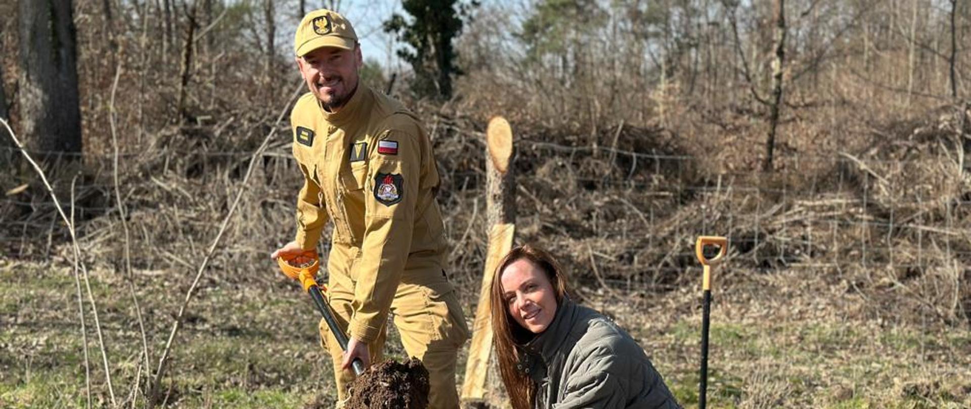 Na zdjęciu funkcjonariusz st. ogn. Zenon Stencel i Pani Anna Pobłocka podczas sadzenia drzewek uśmiechają się do fotografa. Strażak jest ubrany w strój służbowy w kolorze piaskowym, natomiast kobieta w zieloną kurtkę, czarne spodnie oraz czarne buty. Jest piękna słoneczna pogoda oraz bezchmurne niebo. 