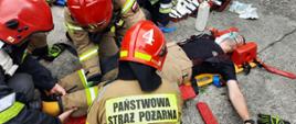 Zdjęcie przedstawia pięciu strażaków podczas ćwiczeń, na desce medycznej leży poszkodowana osoba, którą zaopatrują w pierwszą pomoc. Strażacy ubrani są w ubrania bojowe z hełmem na głowie, obok leży torba medyczna typu R1.