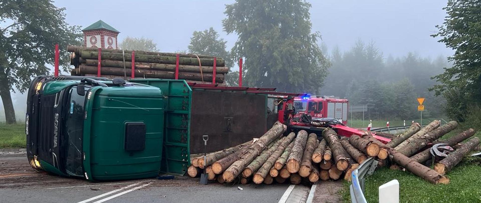 Na jezdni przewrócona ciężarówka przewożąca drzewo. Zielona kabina ciężarówki. Z prawej strony rozrzucone przewożone drzewo. Mgła, w oddali samochody strażackie, zielone drzewa.