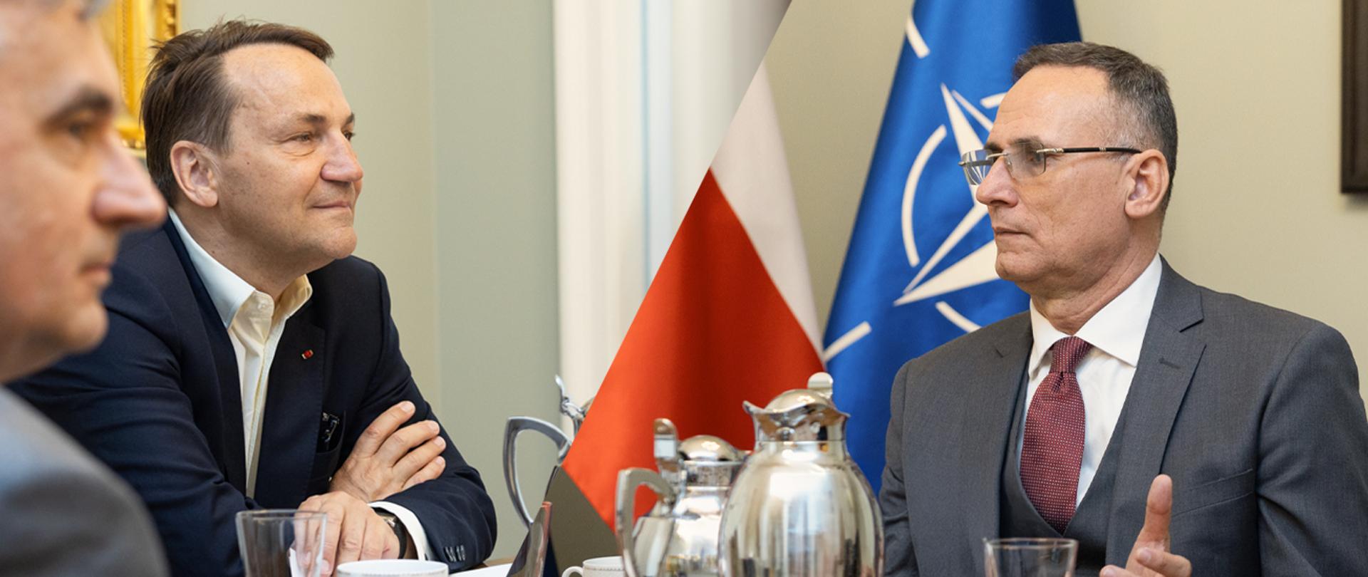 The photo shows two men in suits. They are sitting at a table with glasses and silver pitchers on it. Behind them are flags: the Polish flag and the NATO flag