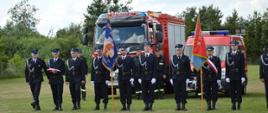 Strażacy ochotniczej straży pożarnej stoją ze sztandarami na tle samochodów gaśniczych.