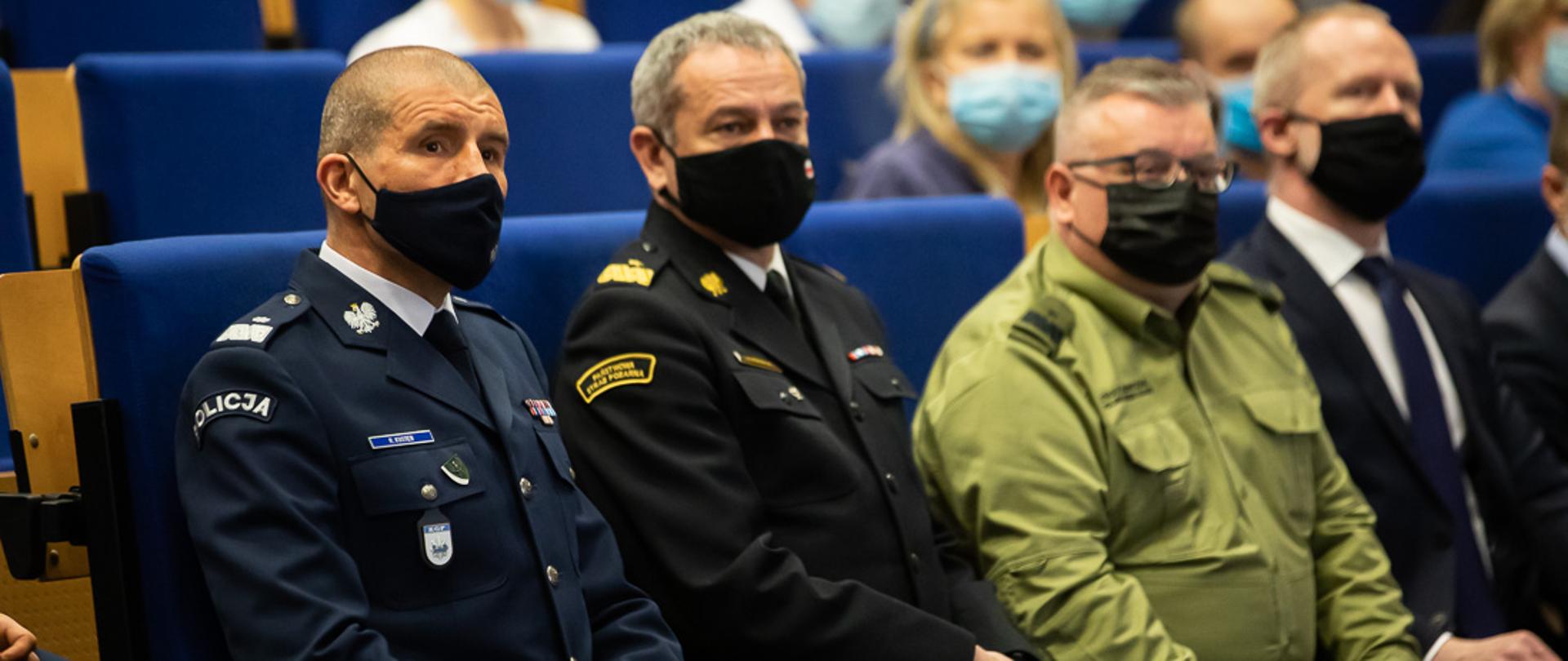 Od lewej, siedzący na sali zastępca komendanta głównego Policji, zastępca komendanta głównego PSP i zastępca komendanta głównego Straży Granicznej. W tle zebrani goście. 